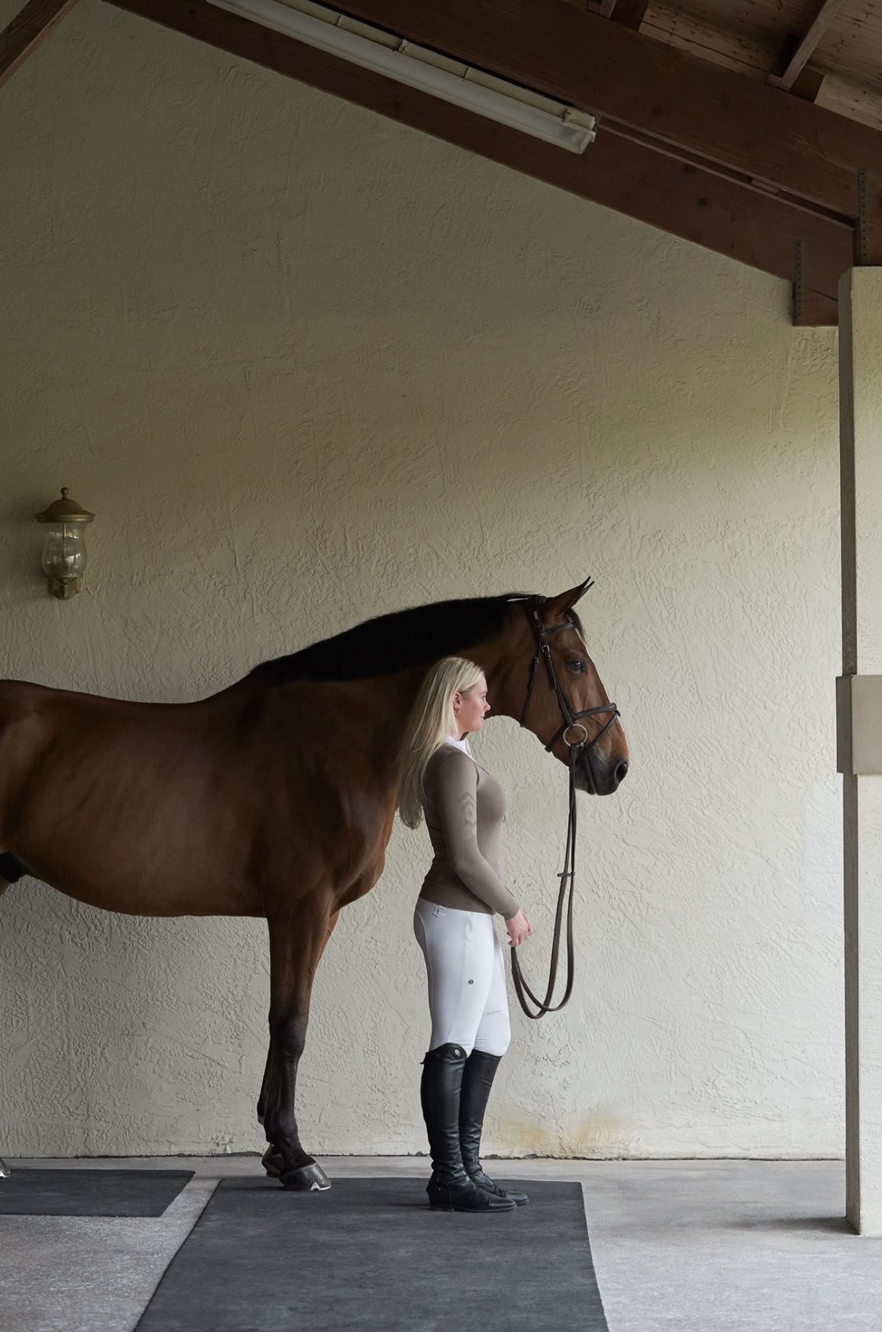 Rider in a taupe top and white breeches standing beside a bay horse under a stable roof